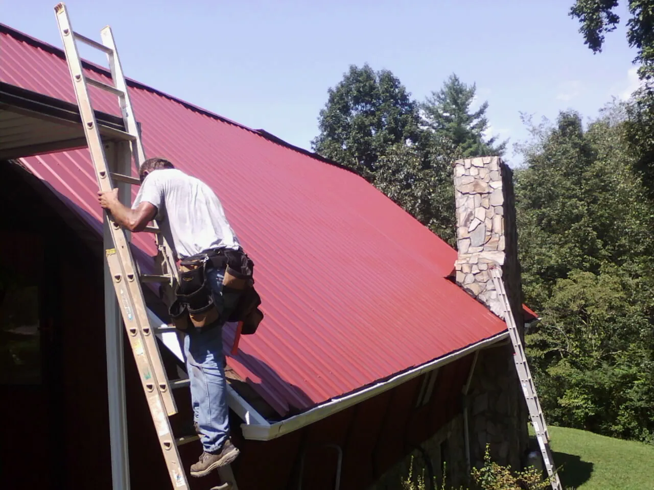 Roofer on a ladder beside a red metal roof and stone chimney — Soffit & Fascia Repair by Roofing Brothers in Leominster
